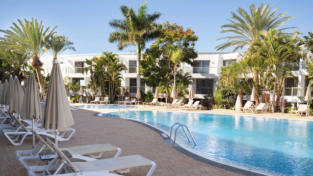 A poolside area with lounge chairs and umbrellas, palm trees, and a building in the background.