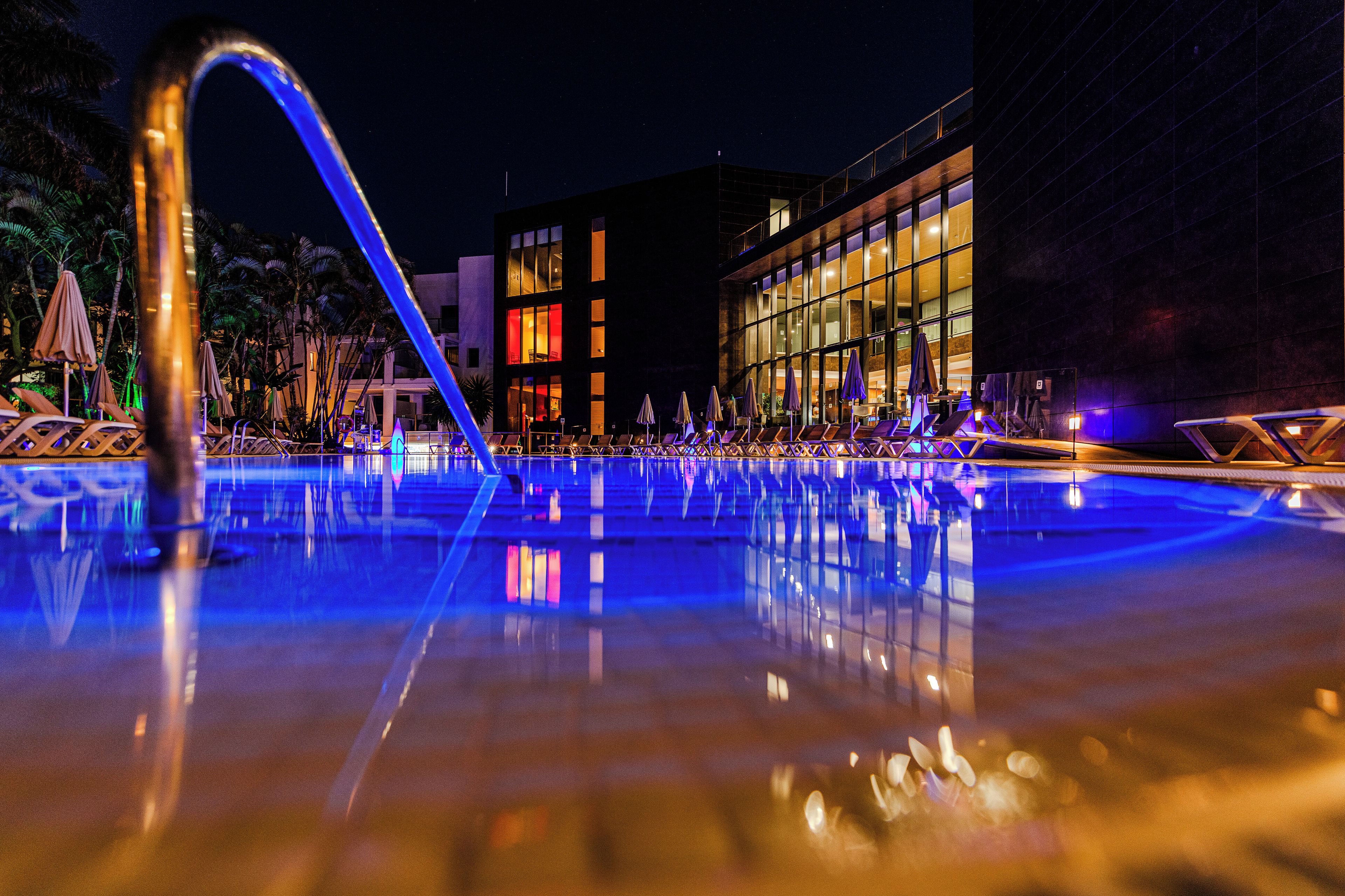 Una piscina nocturna con superficie de agua cristalina que refleja luces, rodeada de sillones de descanso y sombrillas, con un edificio moderno al fondo.