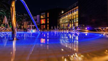 Una piscina nocturna con superficie de agua cristalina que refleja luces, rodeada de sillones de descanso y sombrillas, con un edificio moderno al fondo.