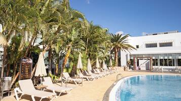 Zona de piscina con sillas de descanso y sombrillas, palmeras y un edificio con un cartel que dice "Oceania".