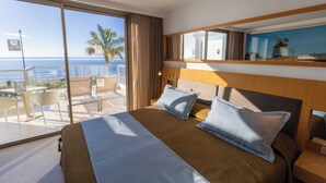 A hotel room with a large bed, two pillows, a nightstand, and a view of the sea through floor-to-ceiling windows.