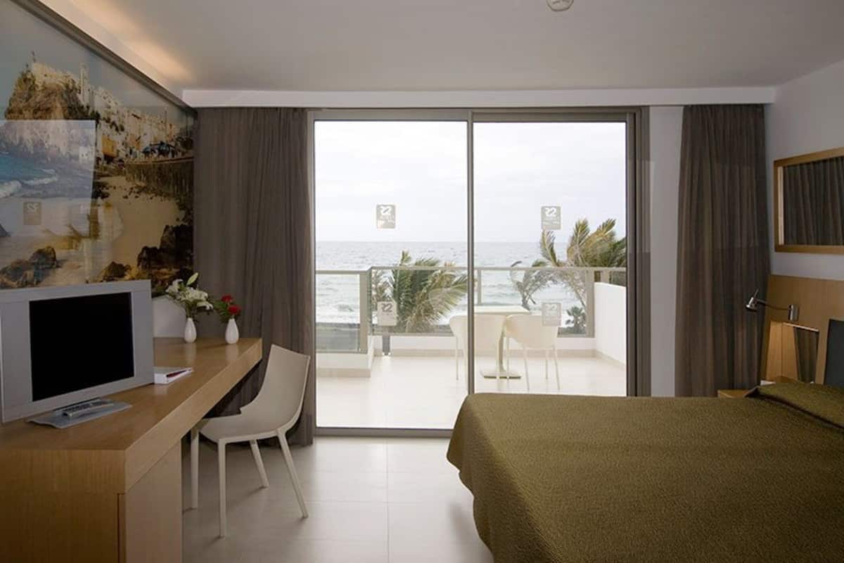 Une chambre d’hôtel avec un lit, un bureau avec une télévision, une chaise, un balcon avec vue sur la mer et un tableau au mur.