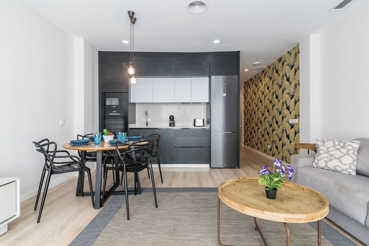 A modern kitchen with a black backsplash, a dining area with a wooden table and black chairs, and a grey sofa with patterned cushions.