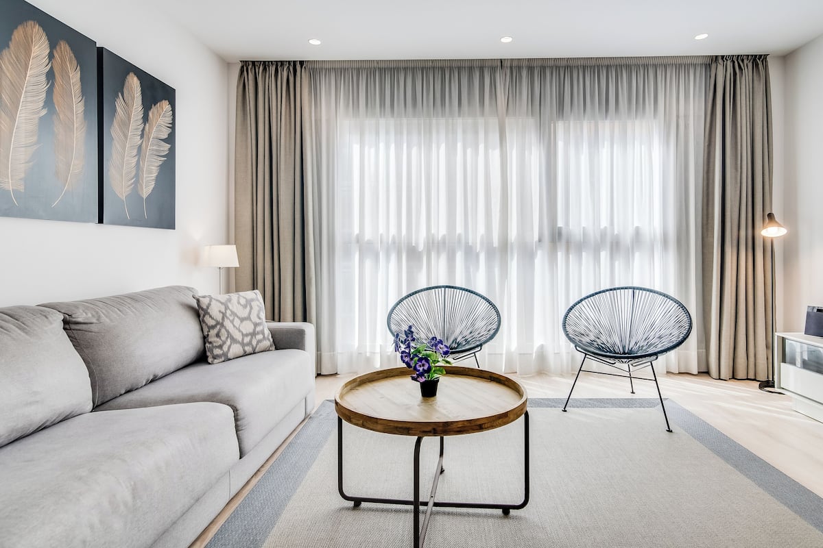 Una sala moderna con un sofá gris, dos sillas de estructura metálica negra, una mesa de centro de madera con un jarrón de flores y un ventanal con cortinas translúcidas.