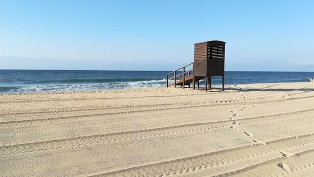 Ein Strand mit einer Holzhütte, möglicherweise einem Rettungsturm, und blauem Himmel.
