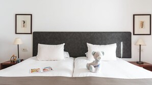 A neatly made bed with white linens, a dark headboard, and a teddy bear on the pillows. There are two framed pictures on the wall, a lamp on each side of the bed, and a magazine on the bed. - Ona Las Lomas Village (Cartagena)