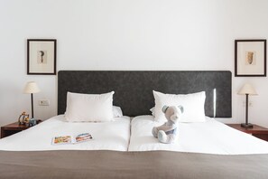 Una cama bien hecha con sábanas blancas, un cabecero oscuro y un osito de peluche sobre las almohadas. Hay dos cuadros en la pared, una lámpara a cada lado de la cama y una revista sobre la cama.