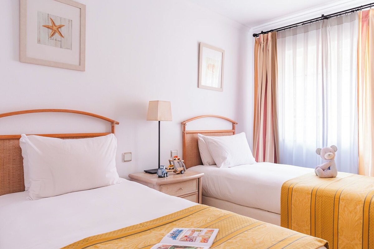 Camera d'albergo con due letti, un comodino con una lampada, un tavolino con un orsacchiotto e una finestra con tende.
