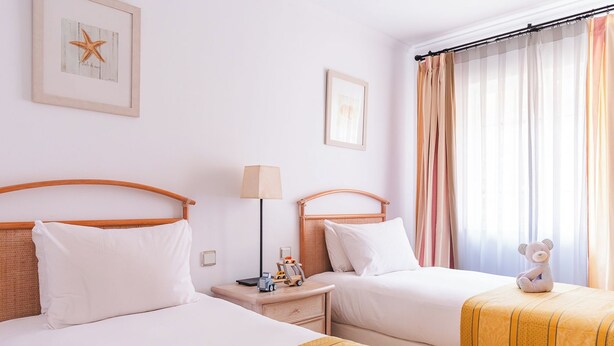A hotel room with two beds, a nightstand with a lamp, a small table with a teddy bear, and a window with curtains. - Ona Las Lomas Village (Cartagena)