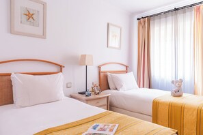 Habitació d'hotel amb dos llits, una taula de nit amb una làmpada, una taula petita amb un os de peluix i una finestra amb cortines.
