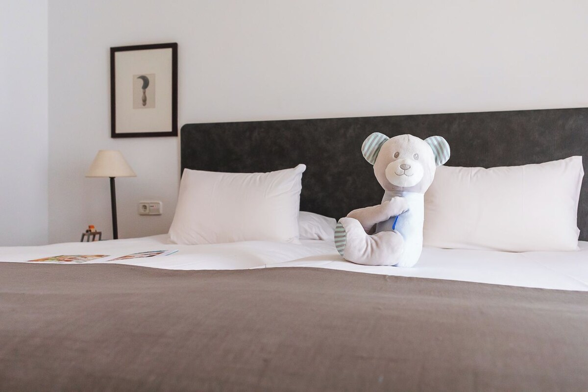 Un letto ben rifatto con lenzuola bianche, un peluche sul letto, un quadro appeso al muro e una lampada accanto al letto.