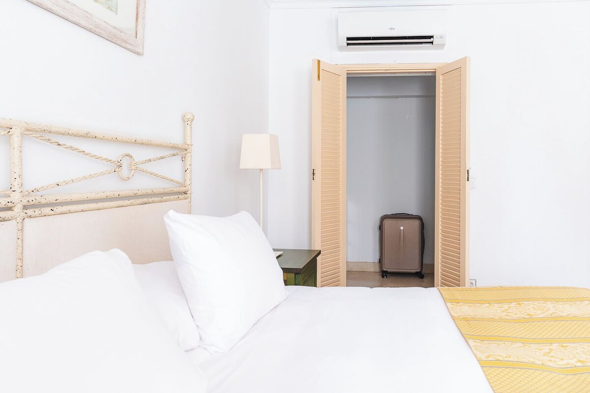 Camera d'albergo con un letto, una testiera in legno, una valigia e una porta aperta che dà su un corridoio.