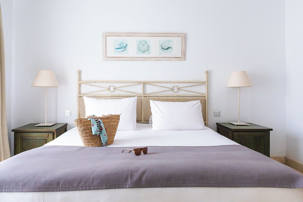 Un letto rifatto con cura, una testiera, due comodini e un quadro incorniciato sopra il letto.