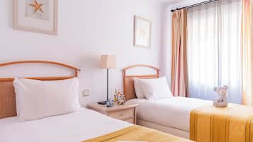 Een hotelkamer met twee bedden, een nachtkastje met een lamp, een kleine tafel met een teddybeer, en een raam met gordijnen.