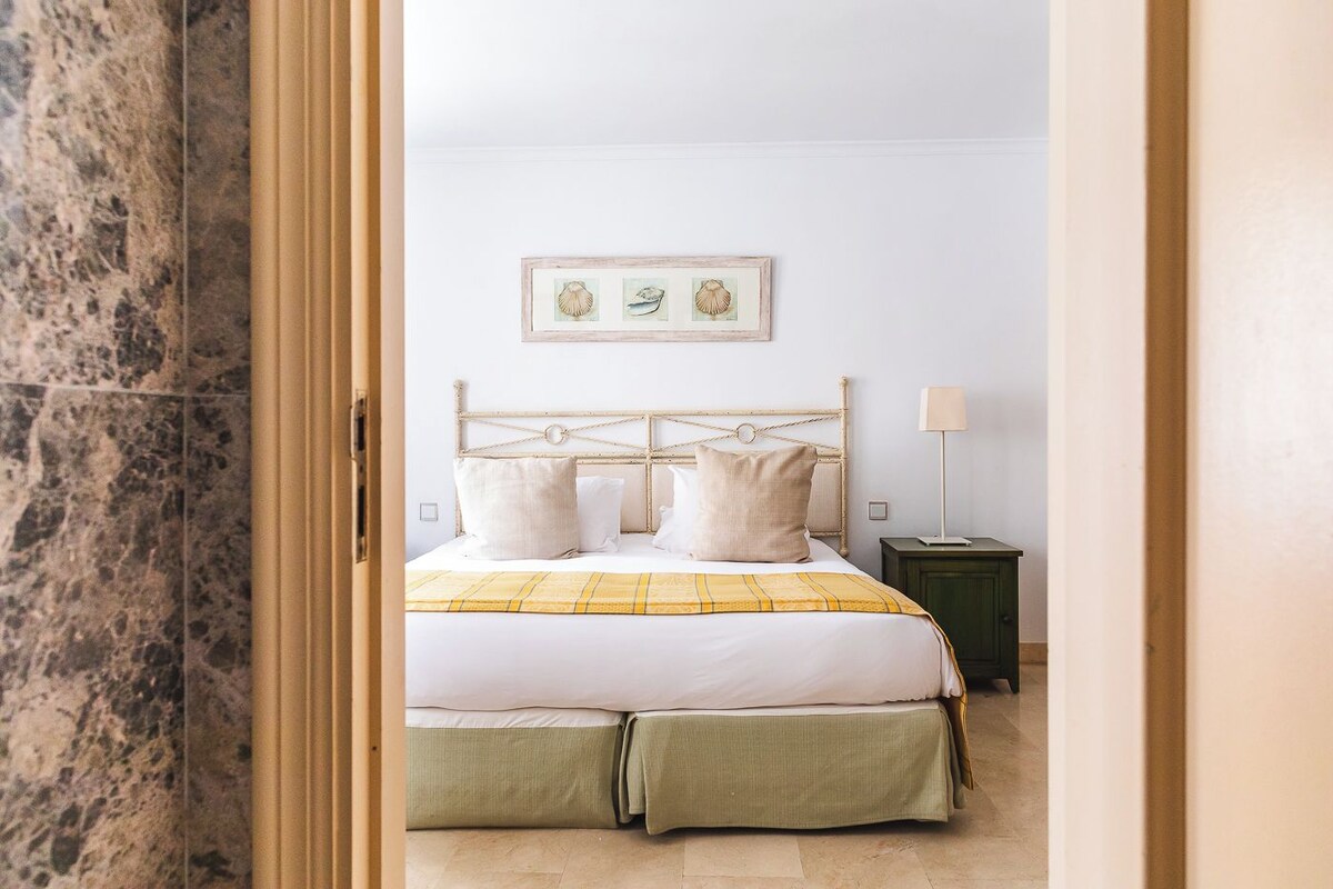 Una camera da letto con un letto, cuscini, un comodino, una lampada e un quadro appeso al muro.
