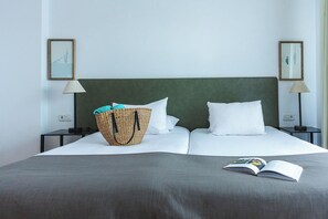 Una cama bien hecha con almohadas blancas y una colcha gris, una cesta de mimbre con toallas y un libro sobre la cama.