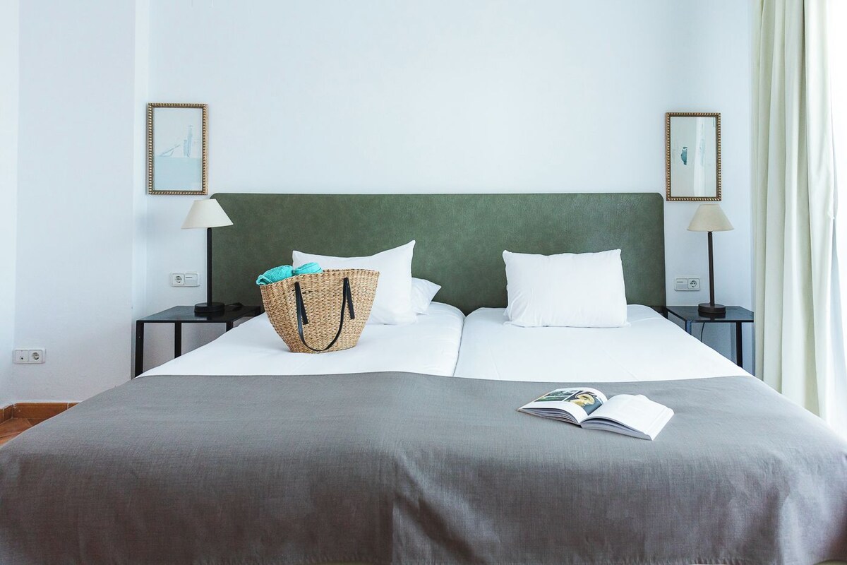 Un letto rifatto con ordine, con copripiumino grigio, due cuscini bianchi, un cesto di vimini con un asciugamano color petrolio e una rivista sul letto.