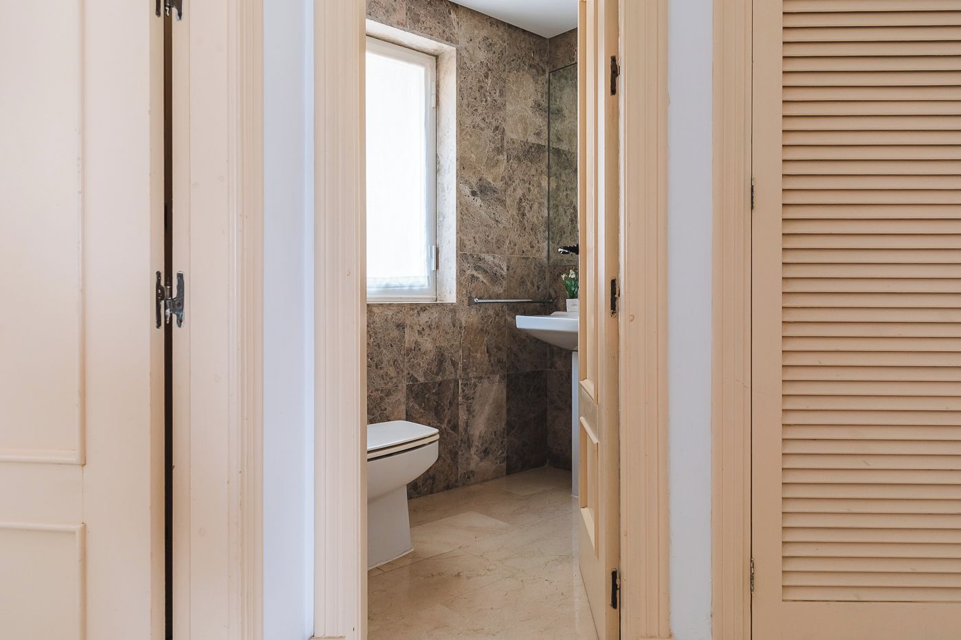 Ein schmaler Flur, der zu einem Badezimmer mit Toilette, Fenster und Waschbecken führt.
