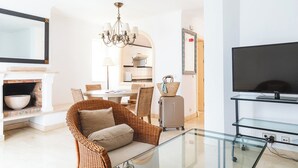 Un salon moderne avec une table basse en verre, des meubles en osier, une cheminée, un coin repas et un téléviseur posé sur un meuble.
