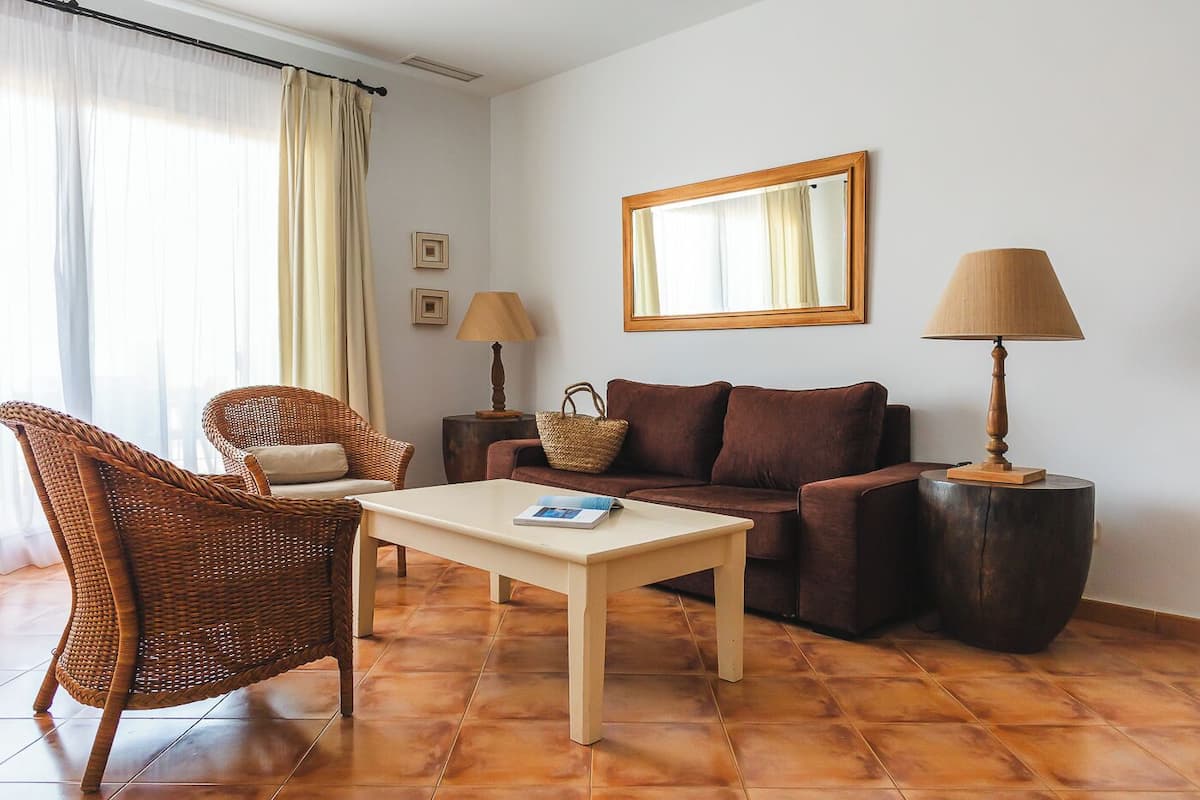 Ein Wohnzimmer mit einer Couch, einem Korbsessel, einem Couchtisch, zwei Lampen und einem Spiegel an der Wand.