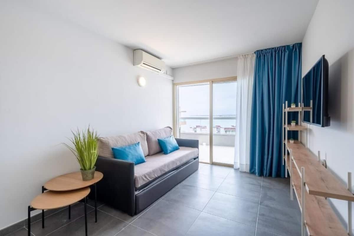 Ein modernes Wohnzimmer mit einer Couch, einem kleinen Tisch mit einer Pflanze, einem an der Wand montierten Fernseher und einem Balkon mit Vorhängen.