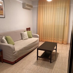Uma sala de estar com um sofá bege, uma cadeira de madeira, uma mesa de centro preta e uma janela grande com cortinas leves.