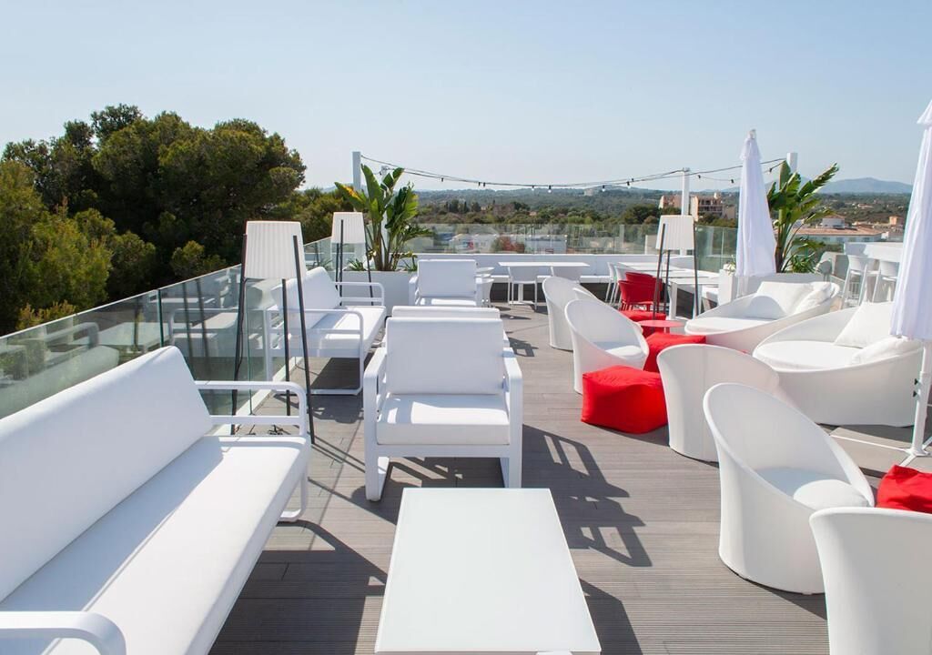 A rooftop terrace with white lounge chairs, red cushions, and a glass railing overlooking a scenic landscape.