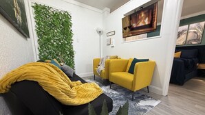 Ein Wohnzimmer mit einer Couch, einem gelben Sessel, einem schwarzen Hocker, einer grünen Wand mit Blätermotiv, einem Flachbildfernseher und einem Bild eines Kamins an der Wand.