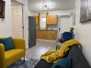 Ein Wohnzimmer mit einer Couch, einem Couchtisch und einem Fernseher. Im Hintergrund ist eine Küche mit Holzschränken und einem Fenster zu sehen.