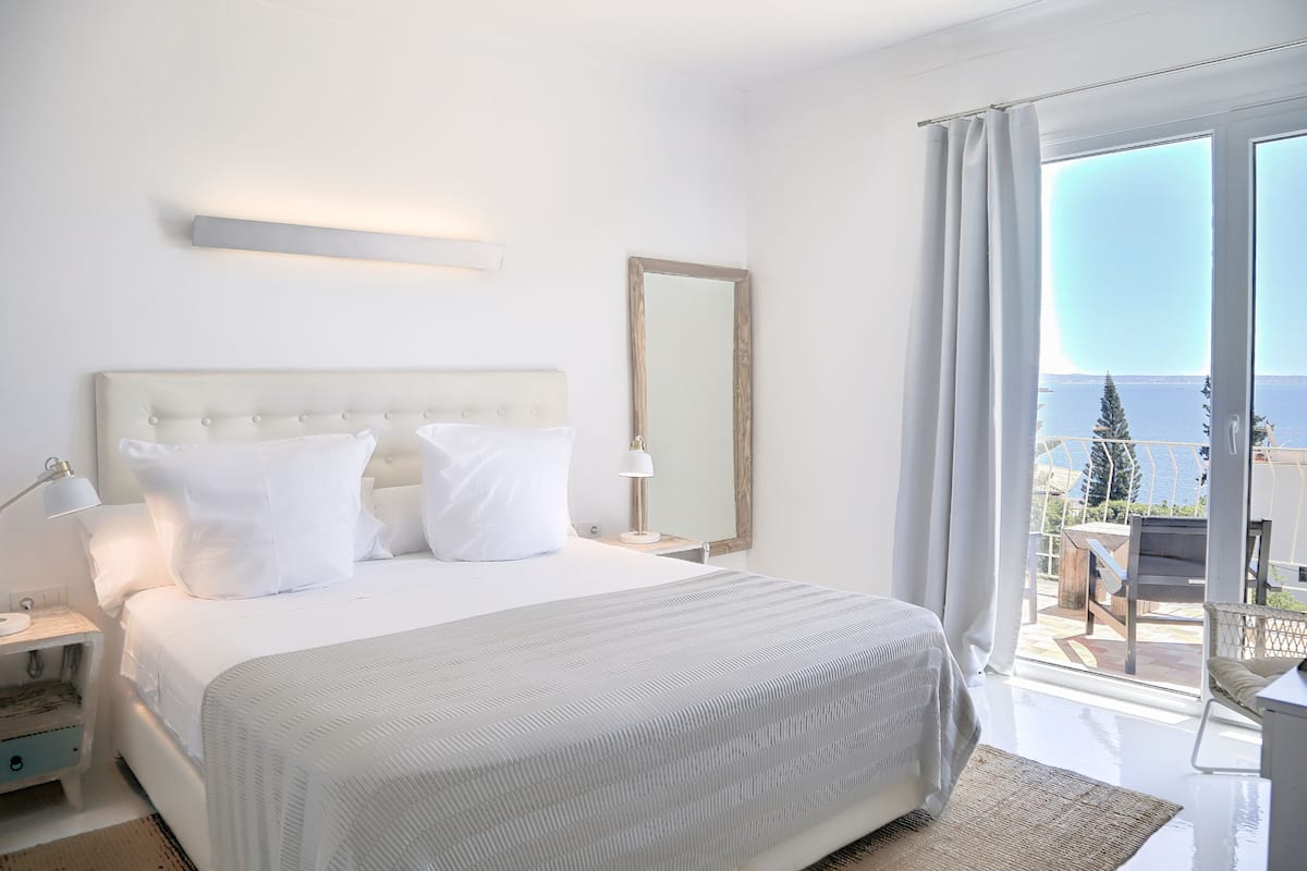 A bedroom with a large bed, a nightstand, a bedside lamp, a mirror, and a view of the sea through a balcony.