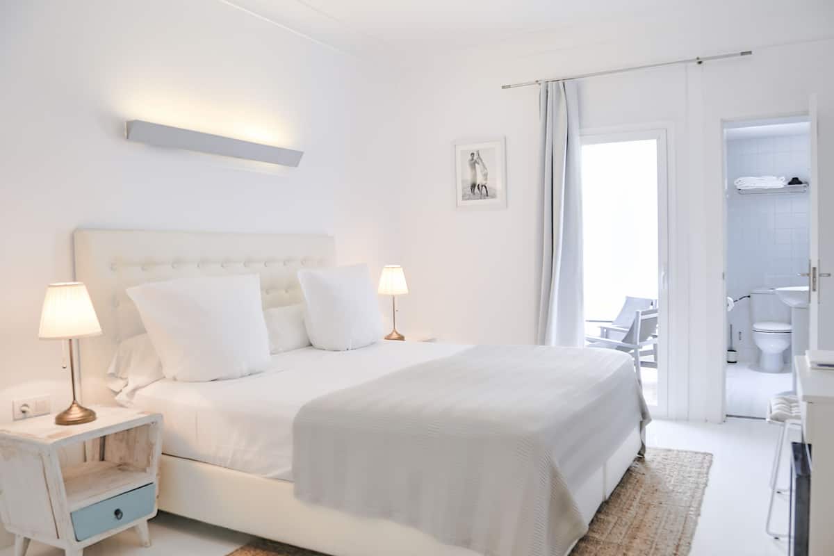 A white bedroom with a large bed, two bedside tables with lamps, a small drawer, a picture on the wall, and an open door leading to a bathroom.