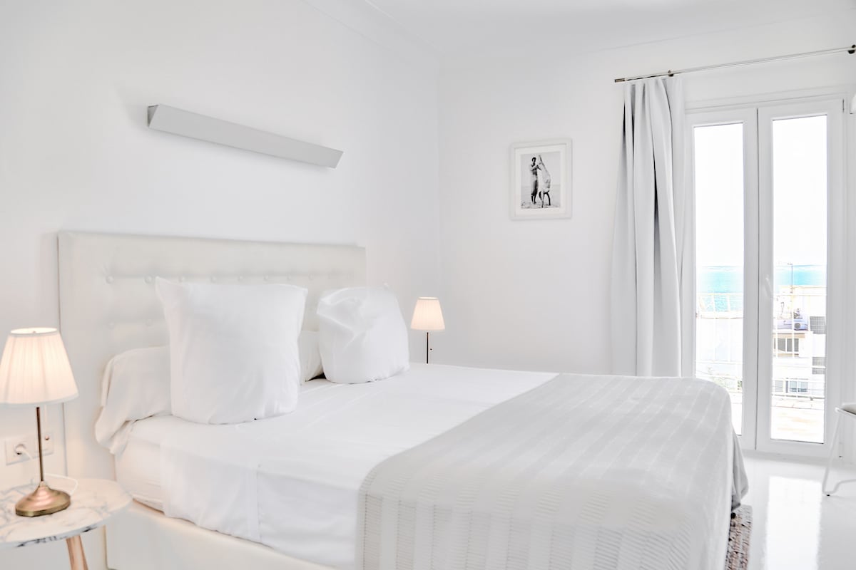 Habitación de hotel con una cama grande, dos lámparas de noche, un cuadro en la pared y vista al mar por la ventana.