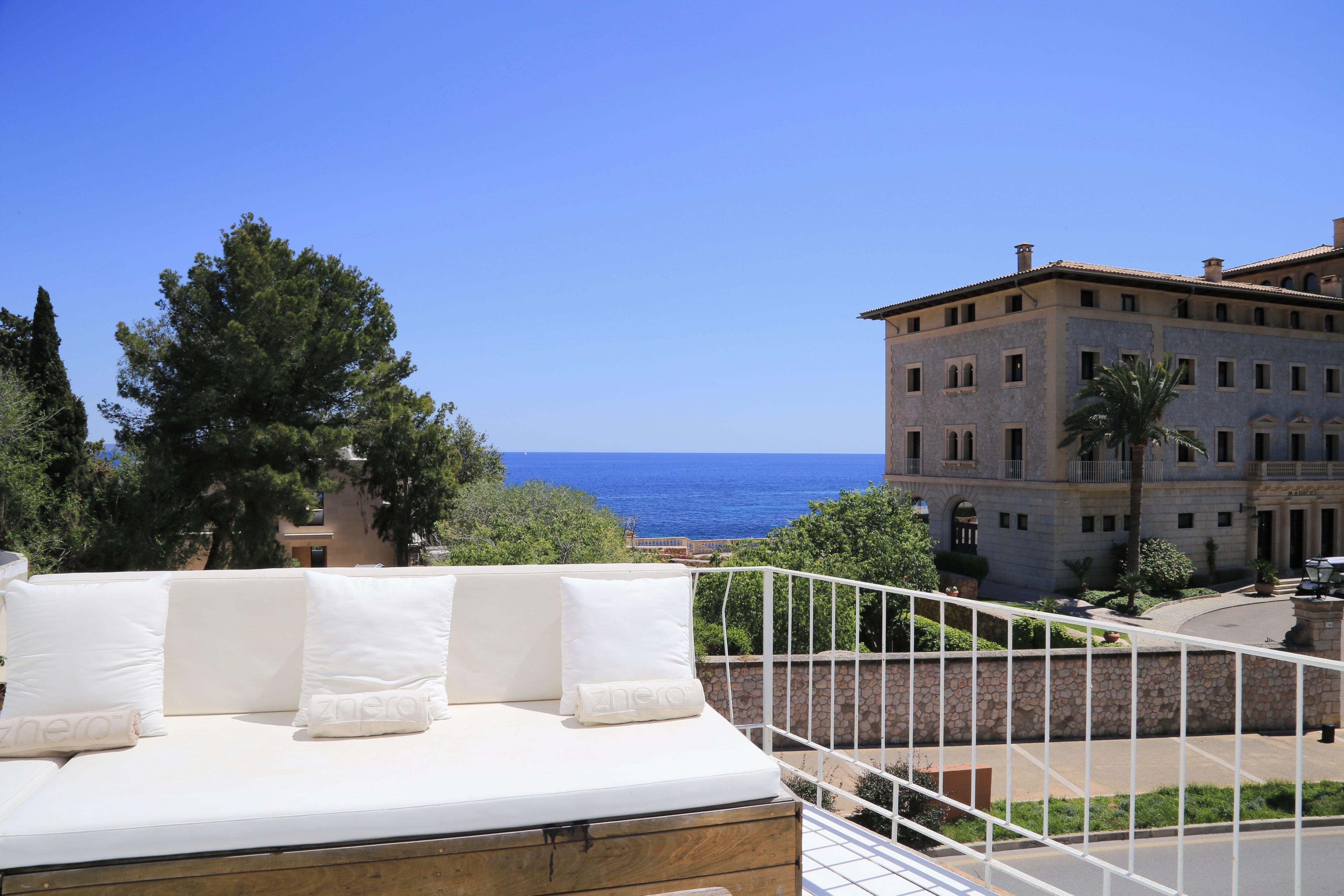 Un balcon avec un canapé blanc et des coussins, donnant sur un immeuble à la façade en pierre et offrant une vue sur la mer.