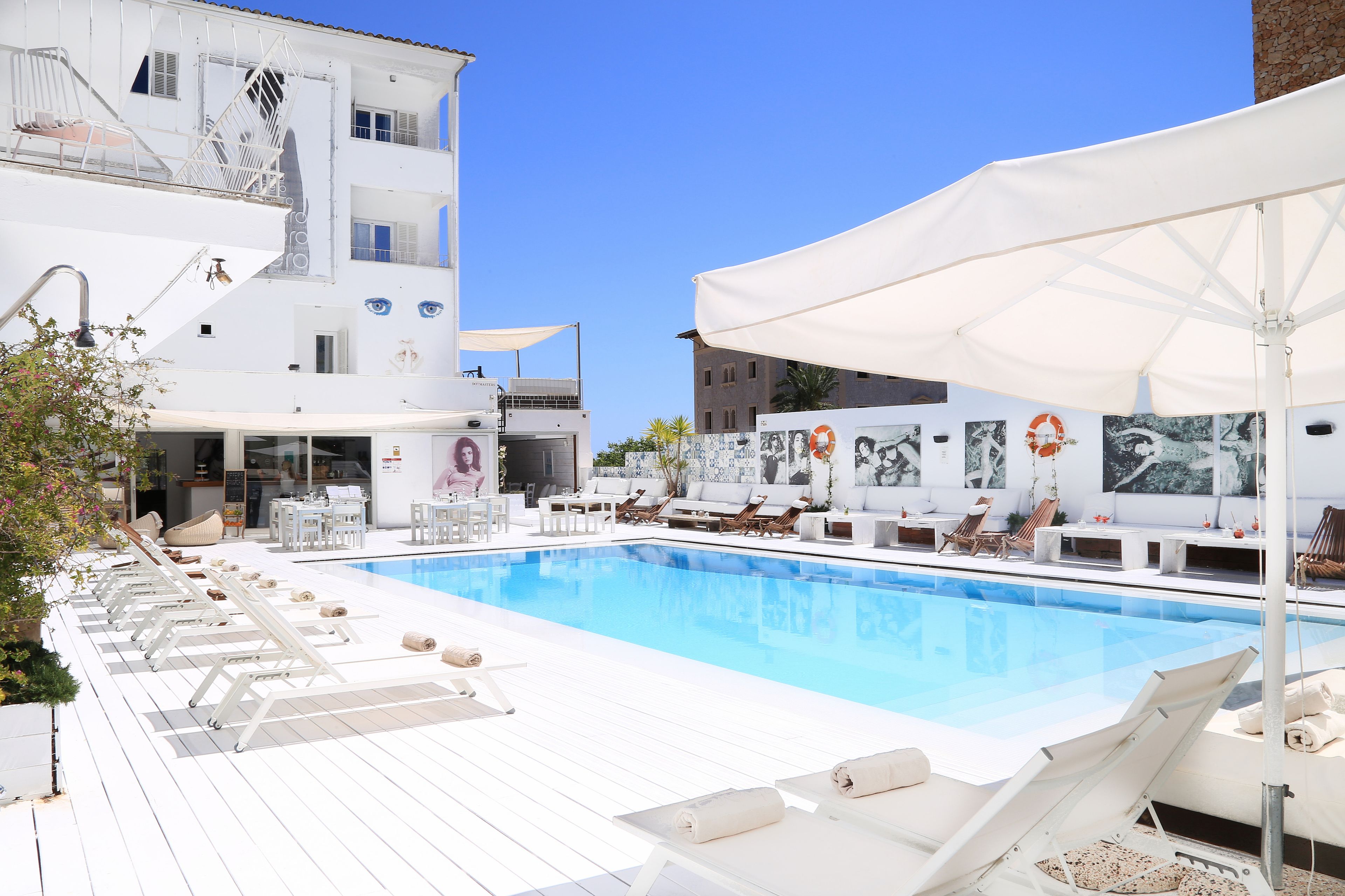 A poolside area with sun loungers, a white umbrella, and a building with a mural in the background.