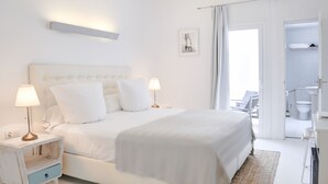 A modern bedroom with a large bed, bedside tables with lamps, a small drawer, a framed picture on the wall, and an open door leading to a bathroom.