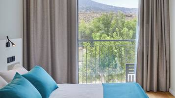 Un dormitorio con un ventanal grande, una cama con ropa de cama azul y blanca, y vistas a un balcón con follaje y colinas.