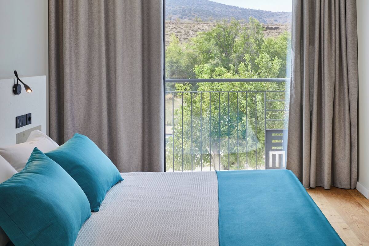 Une chambre avec une grande fenêtre, un lit avec une literie bleue et blanche, et une vue sur un balcon agrémenté de verdure et de collines.