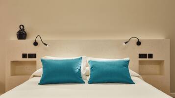 Una cama con dos almohadas color turquesa, una colcha blanca y un cabecero con dos luces fijadas a la pared.
