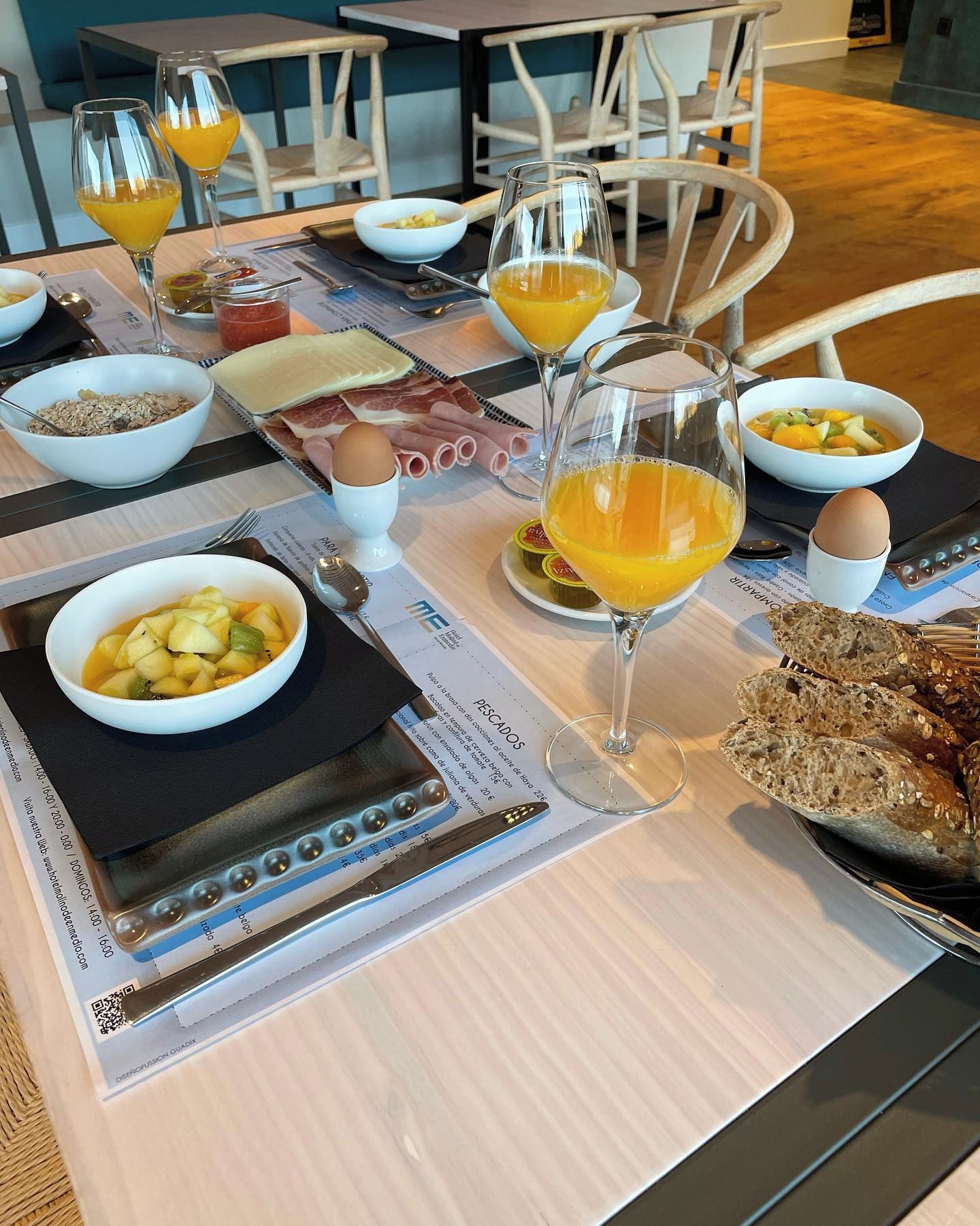 Une table mise pour le petit-déjeuner, avec une nappe pour deux personnes, comprenant des assiettes de nourriture, des verres de jus d’orange et un panier de pain.