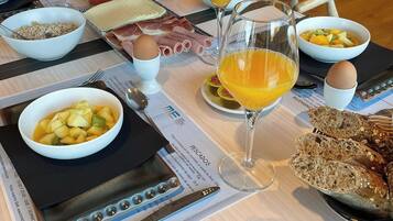 Une table mise pour le petit-déjeuner, avec une nappe pour deux personnes, comprenant des assiettes de nourriture, des verres de jus d’orange et un panier de pain.
