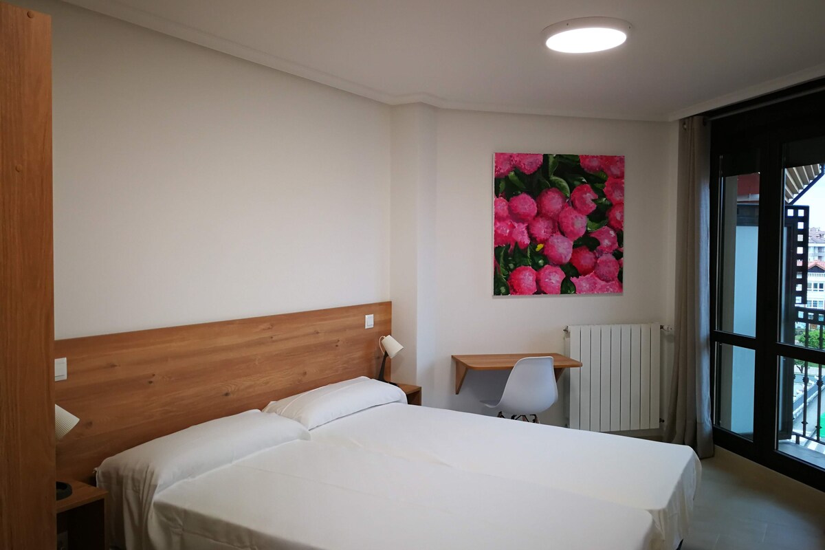 A hotel room with a wooden headboard bed, a small bedside table with a lamp, a radiator, and a window with a view of buildings.