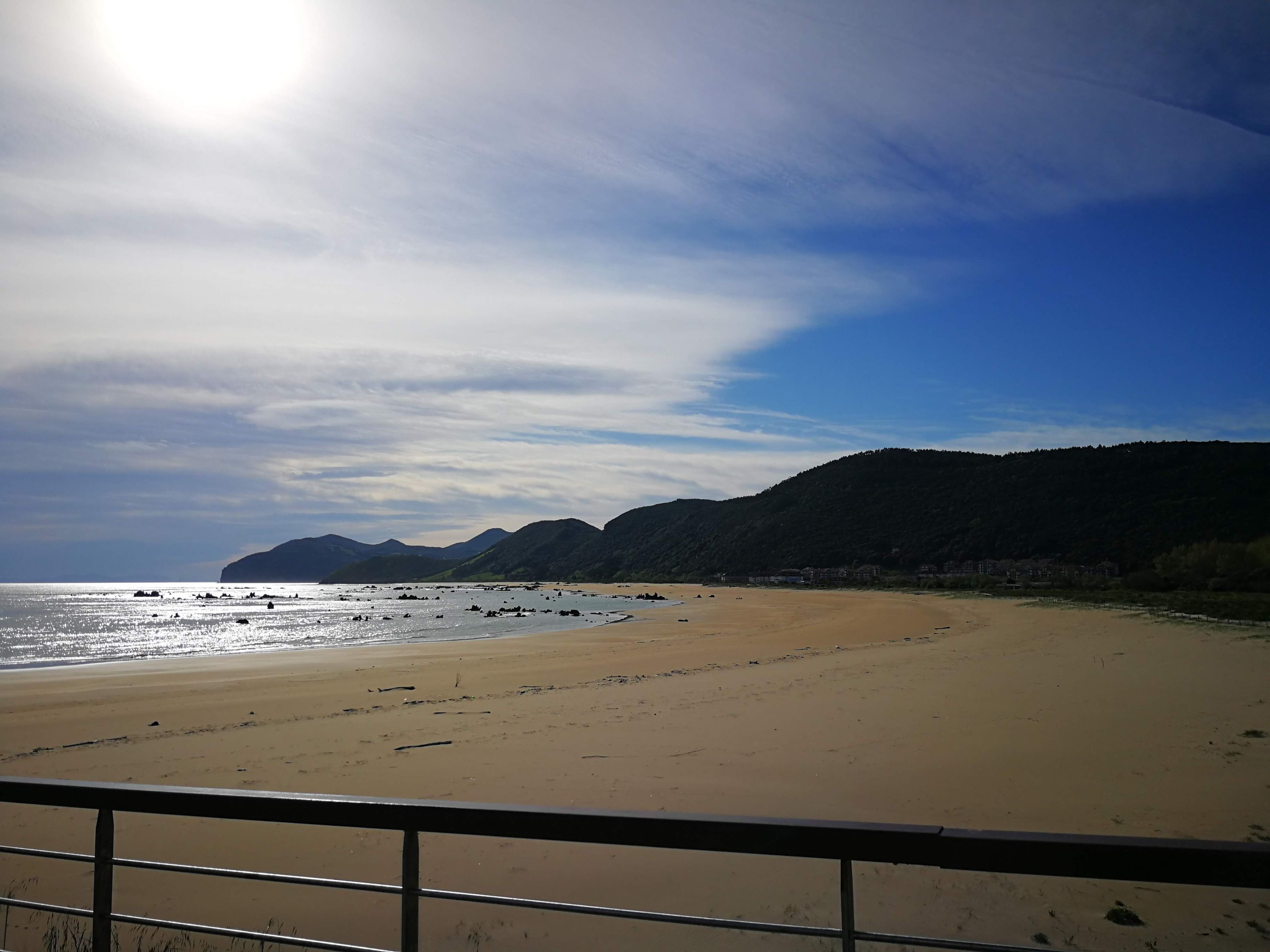 Ein Strand mit blauem Himmel, Bergen im Hintergrund und einem Geländer im Vordergrund.
