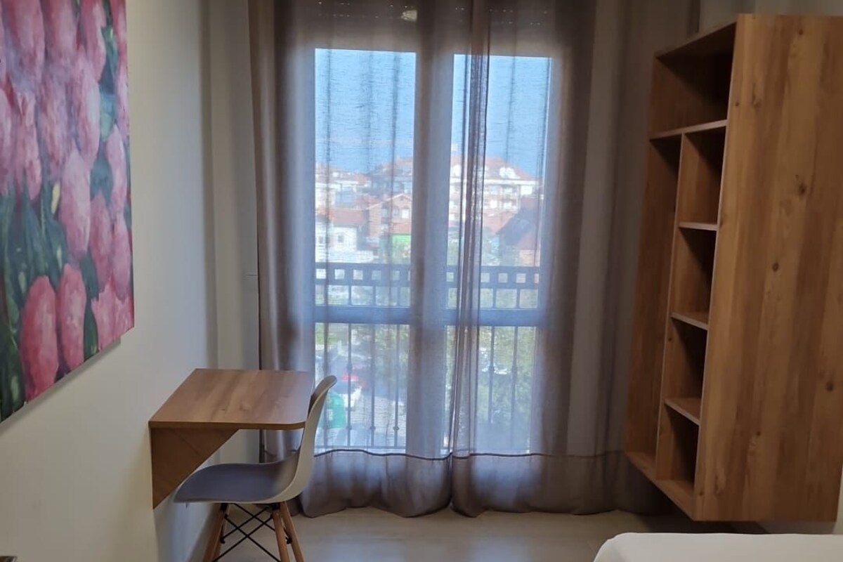 Una habitación con cama, escritorio con silla, estantería de madera, un cuadro en la pared y vistas al exterior.