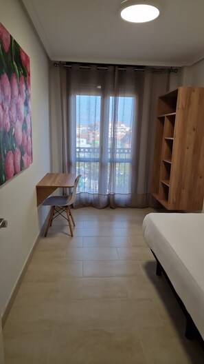 Een kamer met een bed, een bureau met stoel, een houten boekenkast, een schilderij aan de muur en uitzicht door het raam.