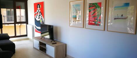 A modern living room with a flat-screen TV on a wooden stand, a sofa, a dining table, and three framed artworks on the wall.