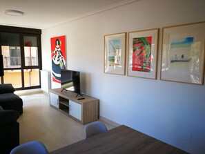 A modern living room with a flat-screen TV on a wooden stand, a sofa, a dining table, and three framed artworks on the wall. - Apartamentos Gran Socaire De Noja (Noja)