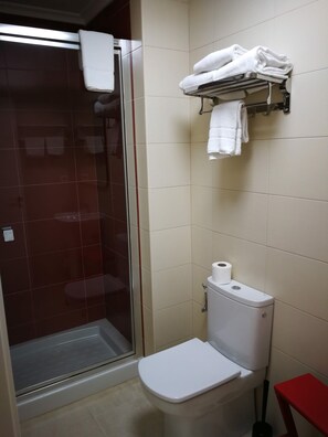 Un baño con cabina de ducha de vidrio, inodoro, un taburete rojo y toallero con toallas dobladas.