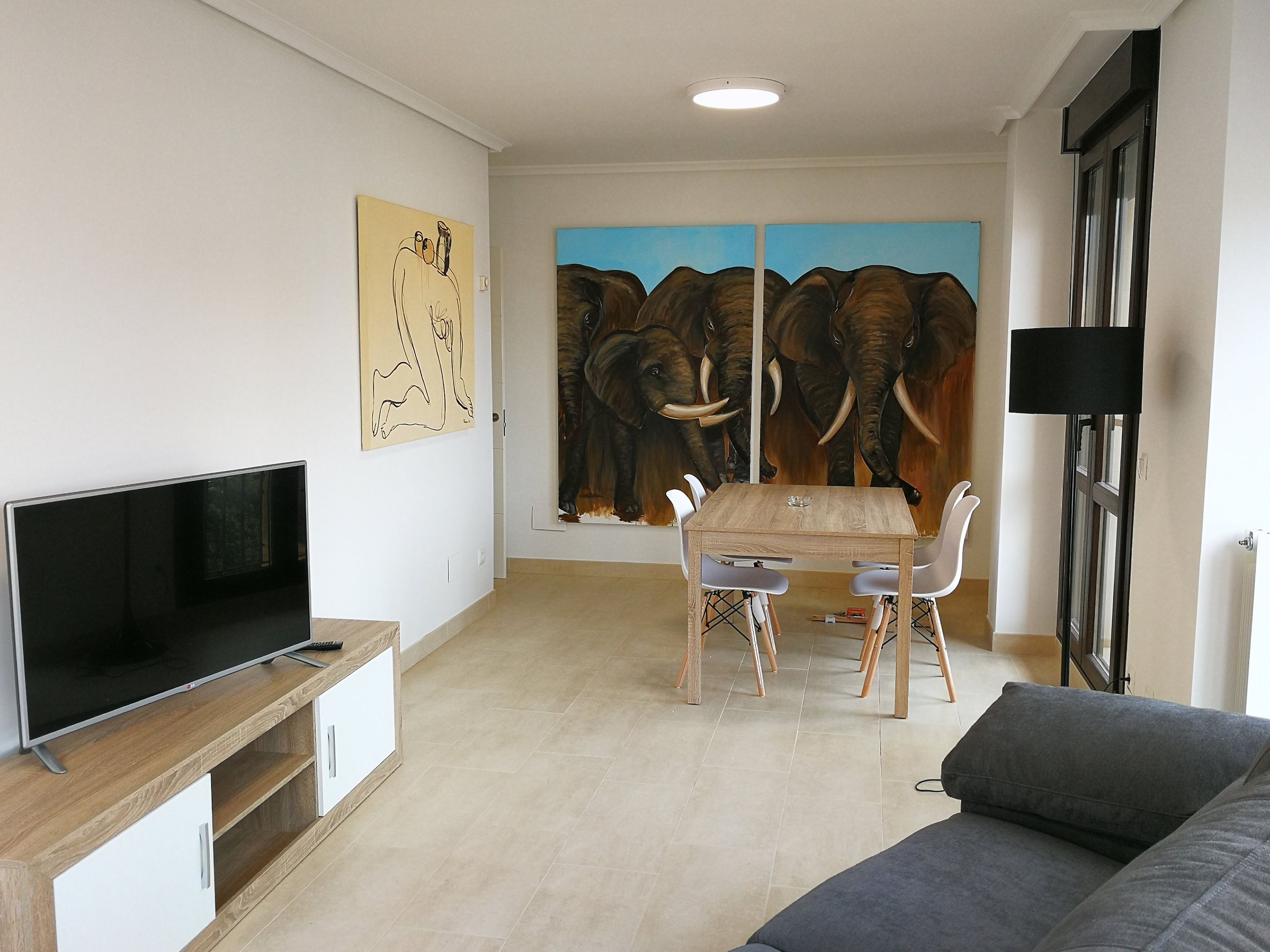 Un salon équipé d’un téléviseur à écran plat, d’une table à manger en bois avec des chaises, et orné d’un grand tableau représentant des éléphants.