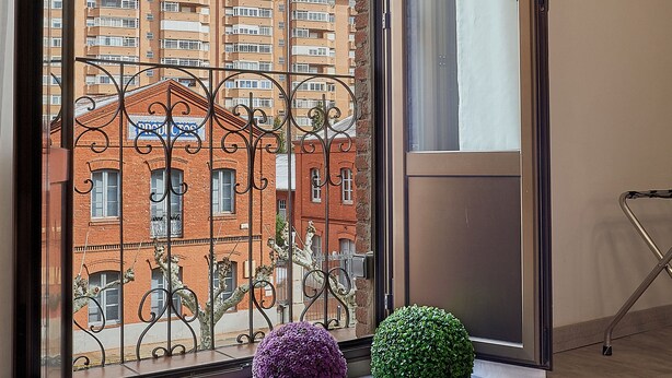 A room with a large window overlooking a residential area, a flat-screen TV mounted on the wall, and two spherical decorative items on the windowsill. - Entrevias Lodging (León)