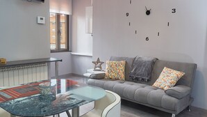 A modern living room with a grey sofa, patterned cushions, a glass coffee table, and a wall clock. - Entrevias Lodging (León)